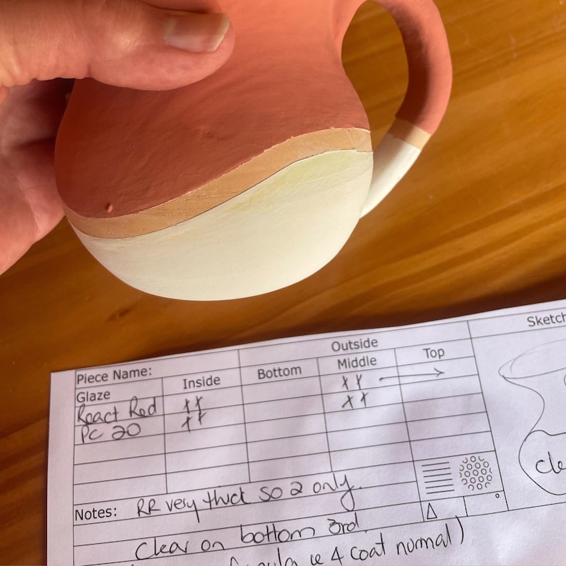 May include: A close-up of a ceramic mug with a red, orange, and white glaze. The mug is labeled "React Red PC 20" and has notes about the glaze being very thick and only two coats being applied.