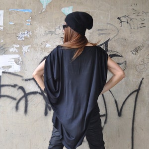 May include: A person wearing a black beanie, black sunglasses, a black oversized shirt, black pants, and black boots. The person is standing in front of a concrete wall with graffiti.