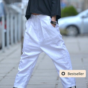 May include: A woman wearing a black top with a geometric necklace and white wide-leg pants. She is wearing sunglasses and has her hair pulled back. The pants have a loose, flowing fit and are cropped at the ankle. The woman is standing on a sidewalk with a car in the background. The image has a "Bestseller" star icon in the bottom right corner.