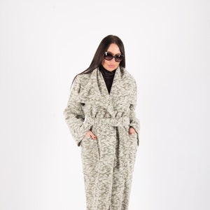 May include: A long, light gray coat with a textured pattern. The coat has a belt at the waist and pockets. The person in the image is wearing sunglasses and black heels. The coat is a stylish outerwear garment.