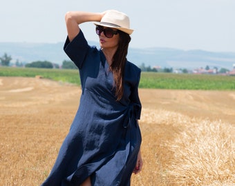 May include: A woman wearing a navy blue linen wrap dress with a white hat and sunglasses, standing in a field of dry grass.