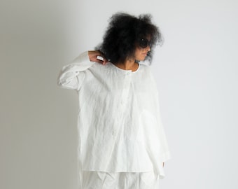 May include: A white long-sleeved button-down shirt with a round neckline and a pair of matching white drop-crotch trousers. The outfit is made of a textured fabric and is paired with black open-toe platform sandals and dark sunglasses.