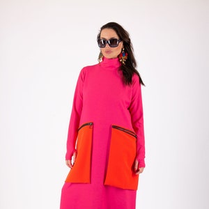 Könnte beinhalten: Ein langes, pinkfarbenes Kleid mit hohem Kragen und zwei großen orangenen Taschen mit schwarzen Reißverschlüssen. Das Kleid ist aus einem weichen, dehnbaren Stoff gefertigt und hat eine lockere Passform. Das Model trägt eine schwarze Sonnenbrille und Ohrringe.