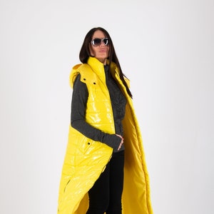 May include: A long yellow puffer vest with a hood. The vest is shiny and has a zipper closure. The model is wearing black pants and black heeled shoes.