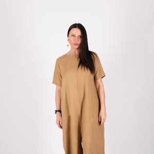 May include: A tan-colored jumpsuit with short sleeves and wide legs. The jumpsuit has a simple, minimalist design with a round neckline. The model is wearing gold earrings and a black watch. The jumpsuit is made of a lightweight fabric.