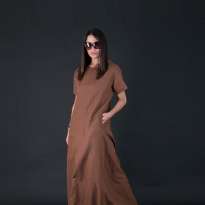 May include: A brown, short-sleeved maxi dress with pockets. The dress is made of a flowing fabric and is worn with black sandals. The model is wearing sunglasses. The dress is a solid color and has a simple, elegant design.