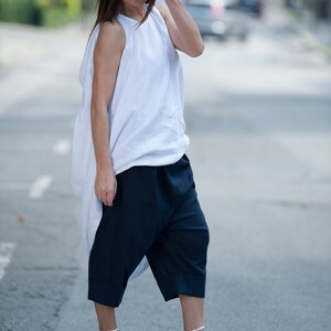 May include: A woman wearing a white, loose-fitting top with a draped neckline and dark blue, wide-legged pants. She is wearing white heeled sandals and sunglasses.