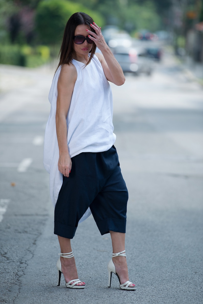 May include: A woman wearing a white, loose-fitting top with a draped neckline and dark blue, wide-legged pants. She is wearing white heeled sandals and sunglasses.