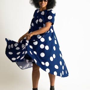 May include: A navy blue dress with large white polka dots. The dress has short sleeves and a flowing, asymmetrical hemline. The person in the image is wearing black high-heeled sandals and sunglasses.
