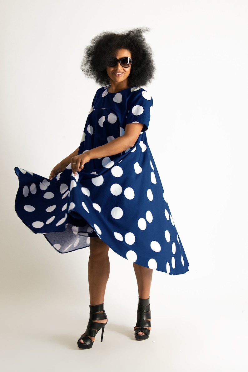 May include: A navy blue dress with large white polka dots. The dress has short sleeves and a flowing, asymmetrical hemline. The person in the image is wearing black high-heeled sandals and sunglasses.