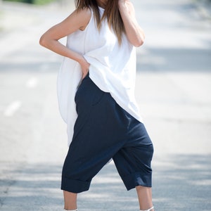 May include: A woman wearing a white sleeveless top with a loose fit and navy blue cropped pants. She is wearing white heeled sandals and sunglasses.