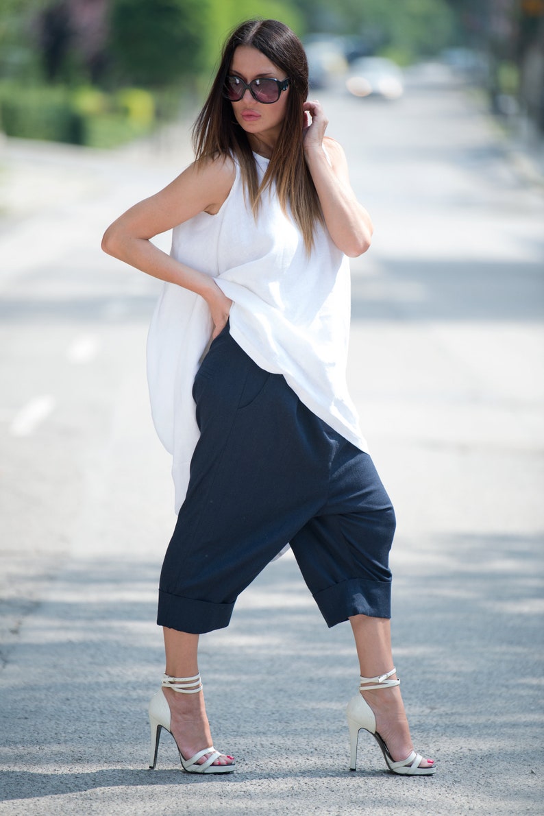 May include: A woman wearing a white sleeveless top with a loose fit and navy blue cropped pants. She is wearing white heeled sandals and sunglasses.