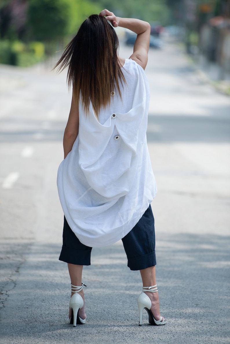 May include: A white linen tunic top with a loose, flowing silhouette. The top features a unique design with a gathered back and a button closure. The model is wearing the top with dark blue cropped pants and white heeled sandals.