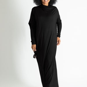 May include: A woman wearing a black maxi dress with a mock neck and long sleeves. She is smiling and holding sunglasses in her hand.