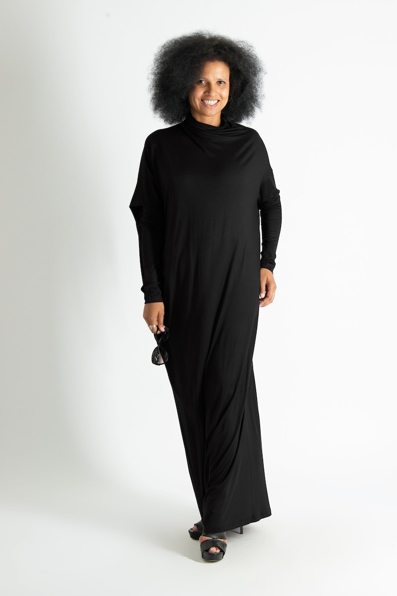 May include: A woman wearing a black maxi dress with a mock neck and long sleeves. She is smiling and holding sunglasses in her hand.