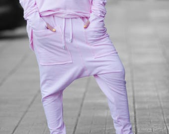 May include: A light pink hoodie and matching drop-crotch pants with pockets. The outfit is paired with red sneakers with white soles and laces. The person is standing on a gray brick surface.