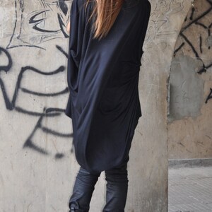 May include: A woman wearing a black long-sleeved tunic top with a black beanie and black boots. The tunic has a loose fit and a cowl neck. The boots have buckles and a chunky heel.