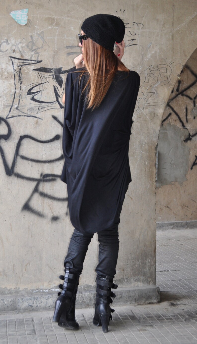 May include: A woman wearing a black long-sleeved tunic top with a black beanie and black boots. The tunic has a loose fit and a cowl neck. The boots have buckles and a chunky heel.