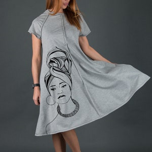 May include: A gray, short-sleeved dress with a black and white illustration of a woman's face on the front. The woman is wearing a headscarf and a necklace.