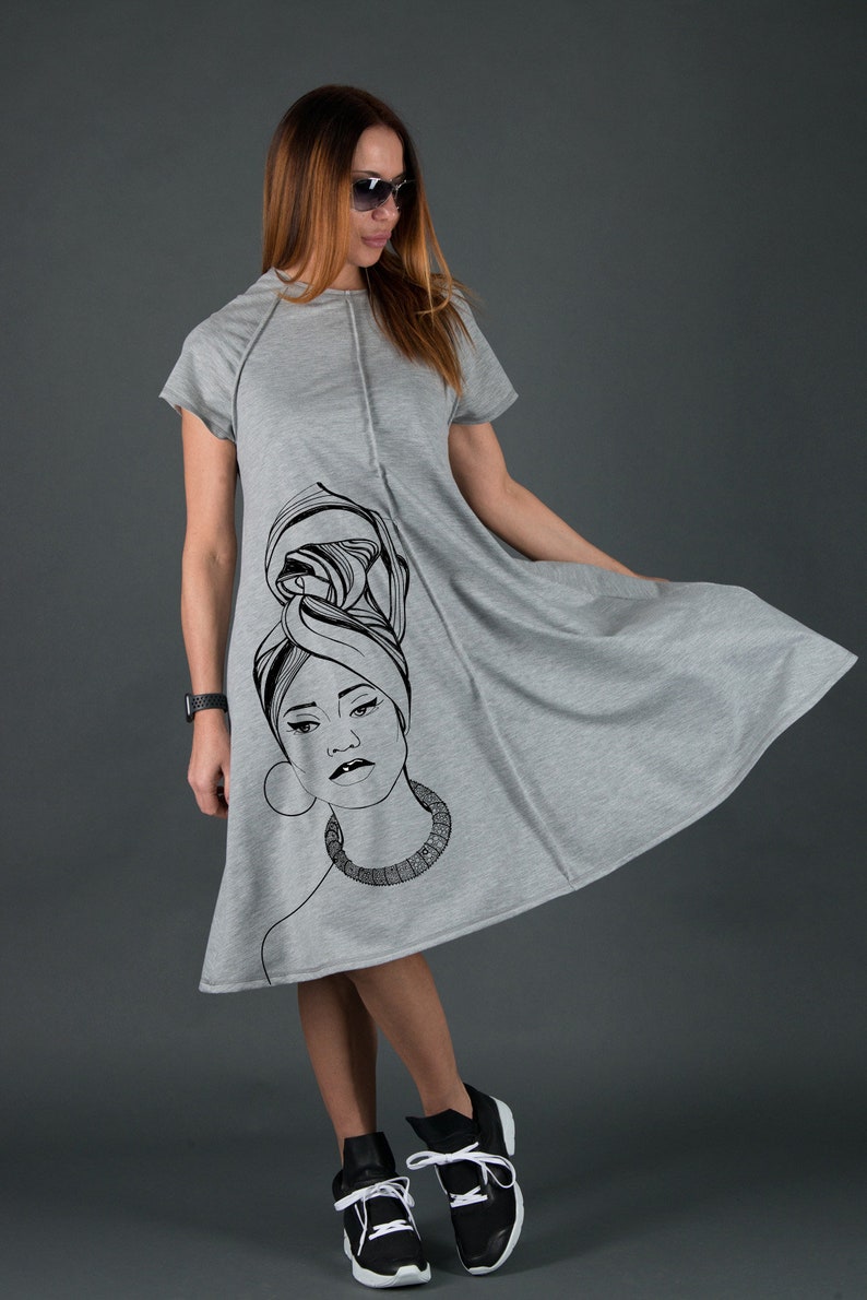 May include: A gray, short-sleeved dress with a black and white illustration of a woman's face on the front. The woman is wearing a headscarf and a necklace.
