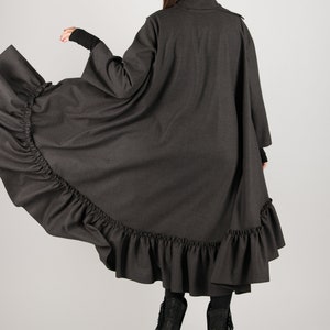 May include: A dark gray, long coat with a ruffled hem. The coat has long sleeves and a high collar. The model is wearing black boots with fringe. The coat is made of a soft, flowing fabric.