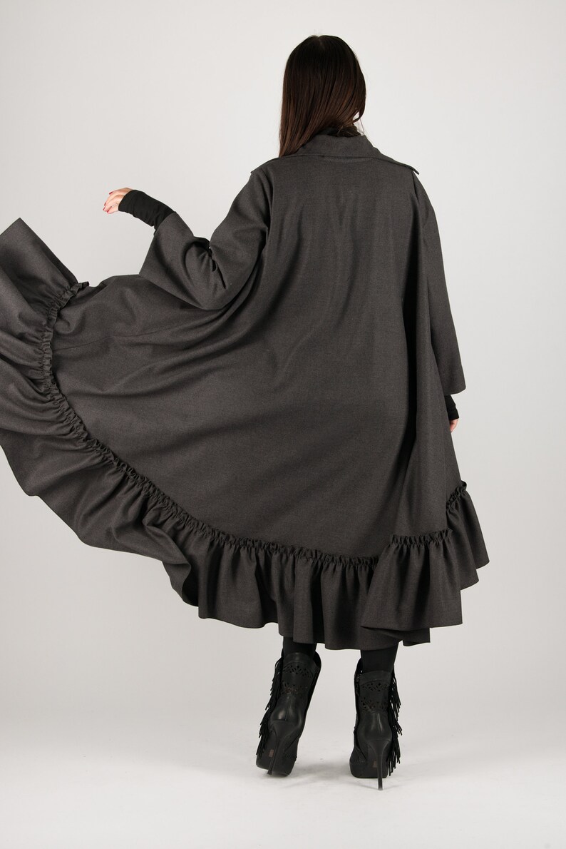 May include: A dark gray, long coat with a ruffled hem. The coat has long sleeves and a high collar. The model is wearing black boots with fringe. The coat is made of a soft, flowing fabric.