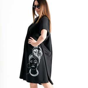 May include: A black short-sleeved dress with a white graphic of a woman's face with a headscarf. The dress has a loose fit and falls to the knees.