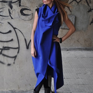 May include: A woman wearing a long, blue, asymmetrical wrap dress with a cowl neck. She is wearing black tights and black heeled boots. The dress has a long, flowing skirt and a fitted bodice.