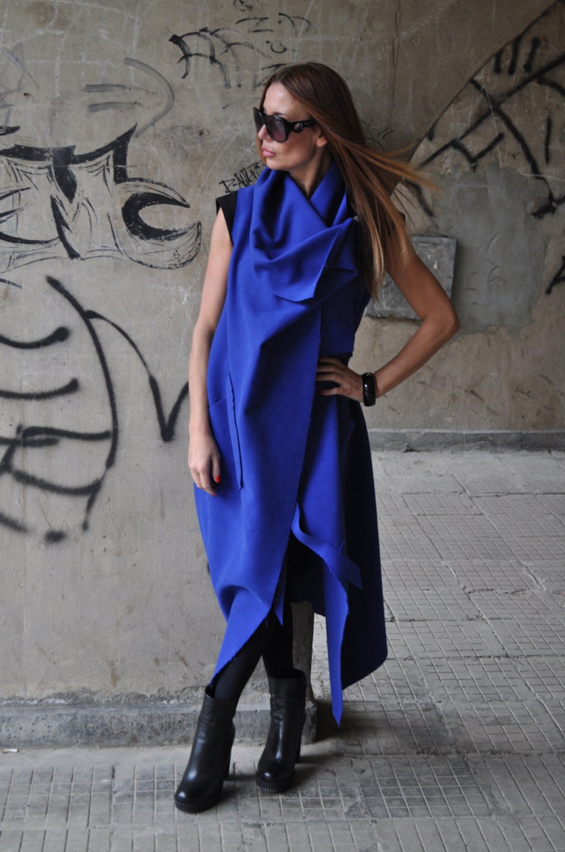 May include: A woman wearing a long, blue, asymmetrical wrap dress with a cowl neck. She is wearing black tights and black heeled boots. The dress has a long, flowing skirt and a fitted bodice.
