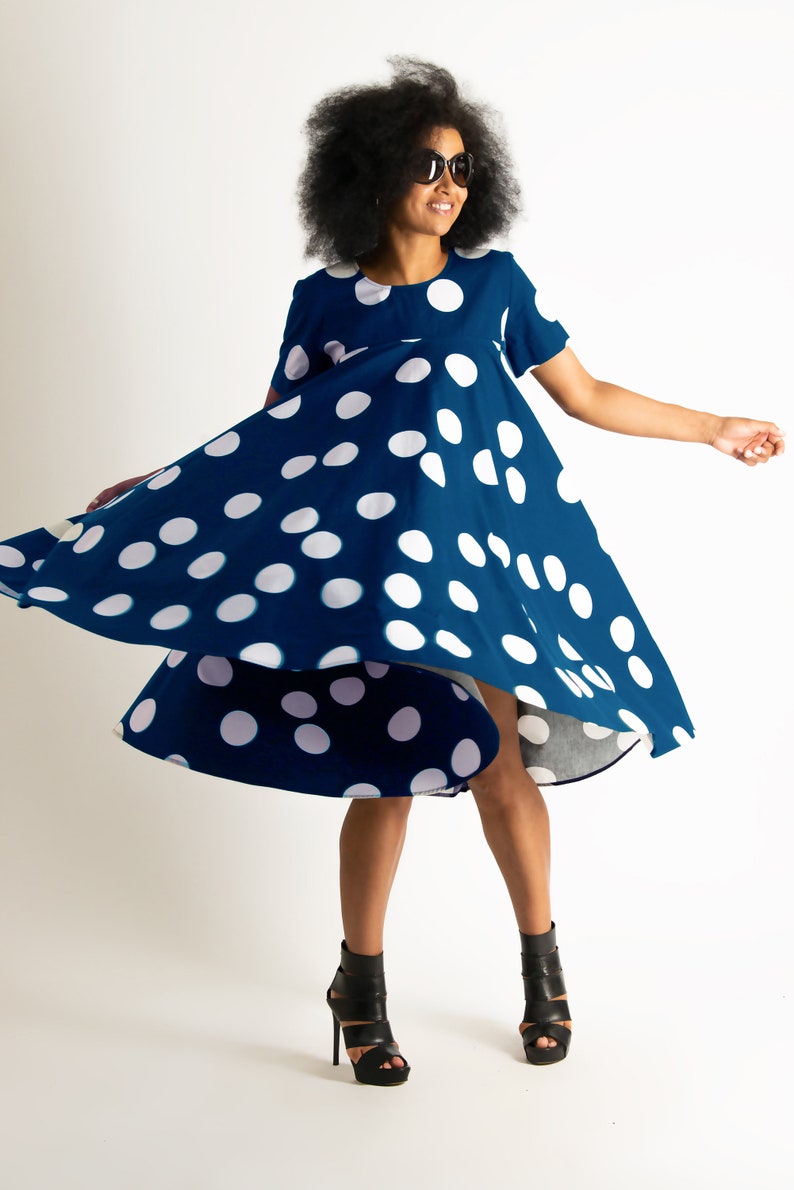 May include: A navy blue dress with large white polka dots. The dress has short sleeves and a flared skirt, and is being twirled. The person in the image is wearing black sunglasses and black high-heeled sandals.