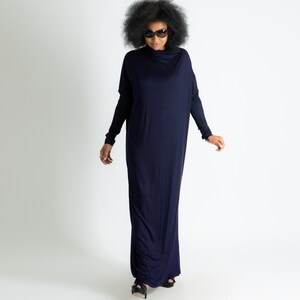 May include: A woman wearing a long, navy blue maxi dress with a cowl neck and long sleeves. She is wearing sunglasses and has her hair in an afro.