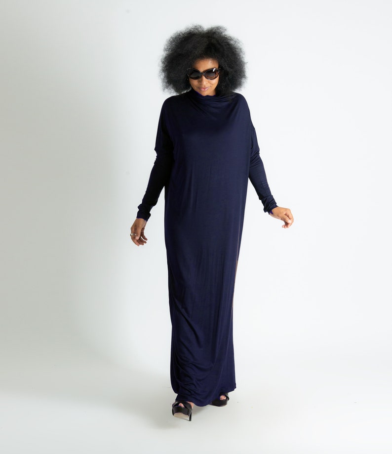 May include: A woman wearing a long, navy blue maxi dress with a cowl neck and long sleeves. She is wearing sunglasses and has her hair in an afro.