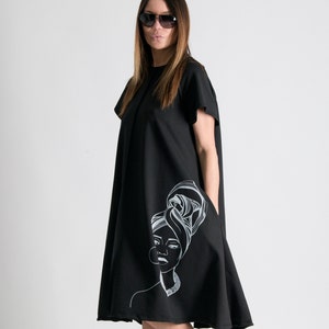 May include: A black midi dress with a white graphic print of a woman with a headscarf. The dress has short sleeves and a loose fit.