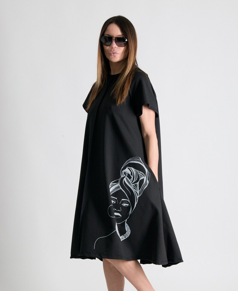 May include: A black midi dress with a white graphic print of a woman with a headscarf. The dress has short sleeves and a loose fit.