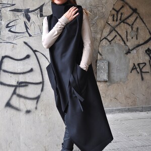 May include: A black asymmetrical long vest with a high collar and pockets. The vest is worn over a beige turtleneck shirt and black pants. The person is wearing black sunglasses and black boots.
