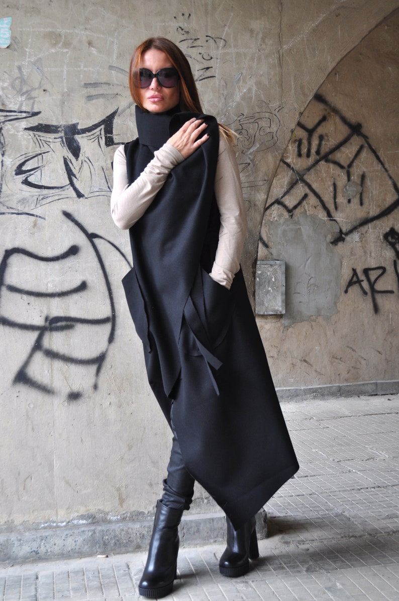 May include: A black asymmetrical long vest with a high collar and pockets. The vest is worn over a beige turtleneck shirt and black pants. The person is wearing black sunglasses and black boots.