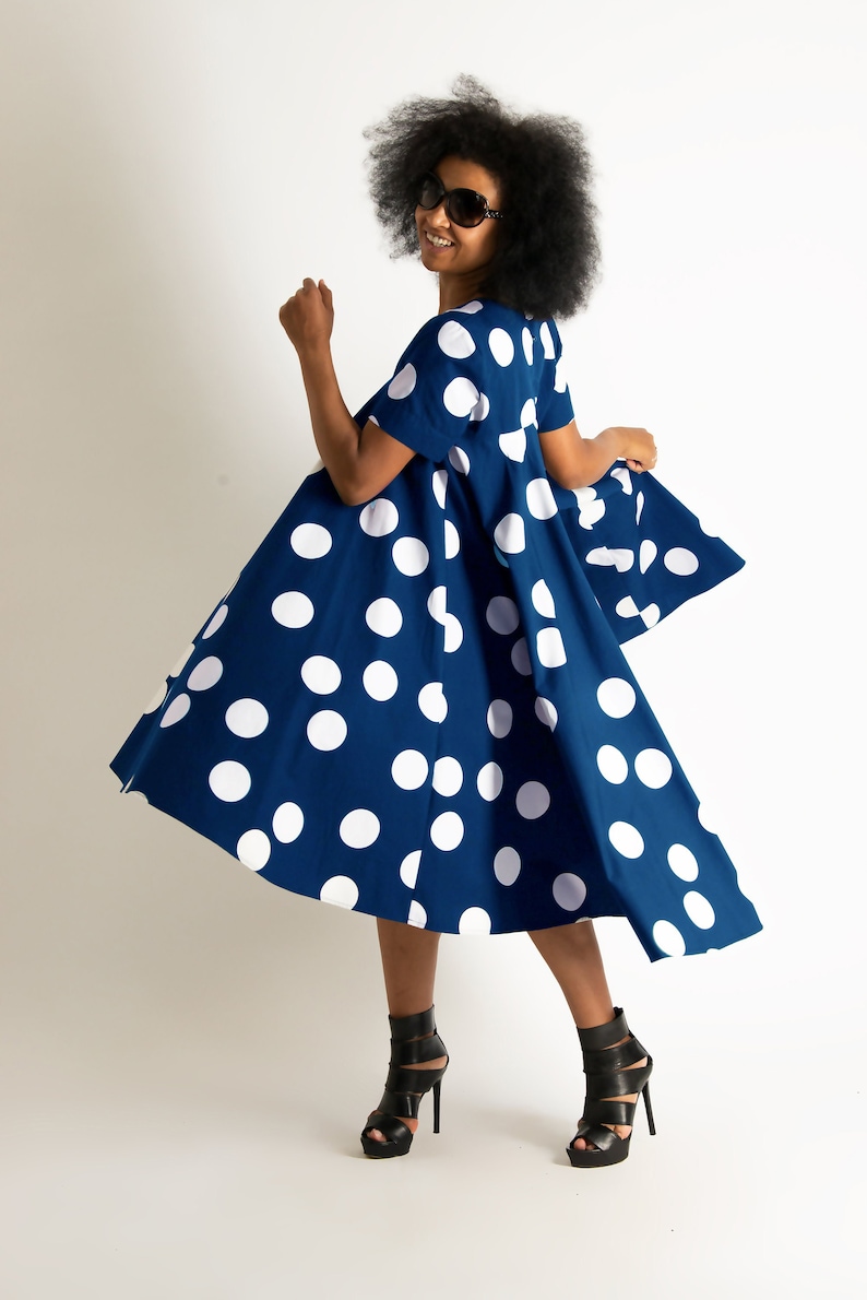 May include: A navy blue dress with large white polka dots. The dress has short sleeves and a flared skirt. The person in the image is wearing black high-heeled sandals and sunglasses. The dress is knee-length.