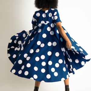 May include: A navy blue dress with large white polka dots. The dress has short sleeves and a full skirt that is in motion. The person is wearing black high-heeled sandals. The dress is a classic, playful style.