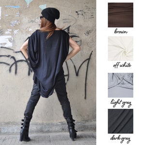 May include: A woman wearing a black oversized top with a draped neckline, black pants, and black ankle boots. She is standing in front of a graffiti-covered wall. The image also shows swatches of fabric in brown, off-white, light grey, and dark grey.