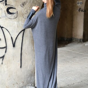 May include: A woman wearing a long, gray, loose-fitting dress with long sleeves. She is standing in front of a wall with graffiti.