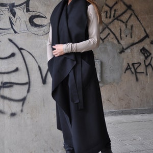 May include: A black long vest with a draped design and a tie belt. The vest is worn over a beige top and black boots.