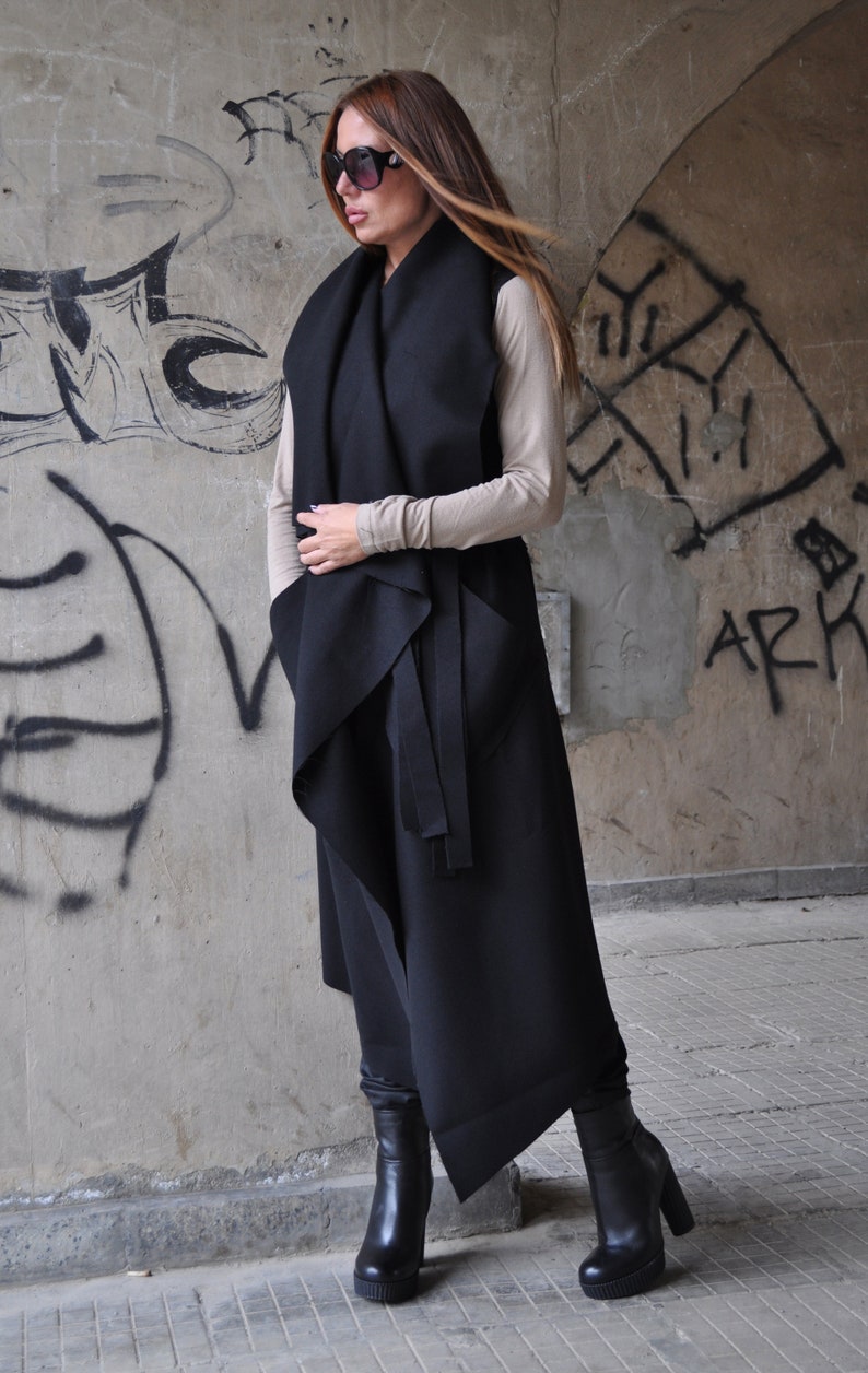 May include: A black long vest with a draped design and a tie belt. The vest is worn over a beige top and black boots.