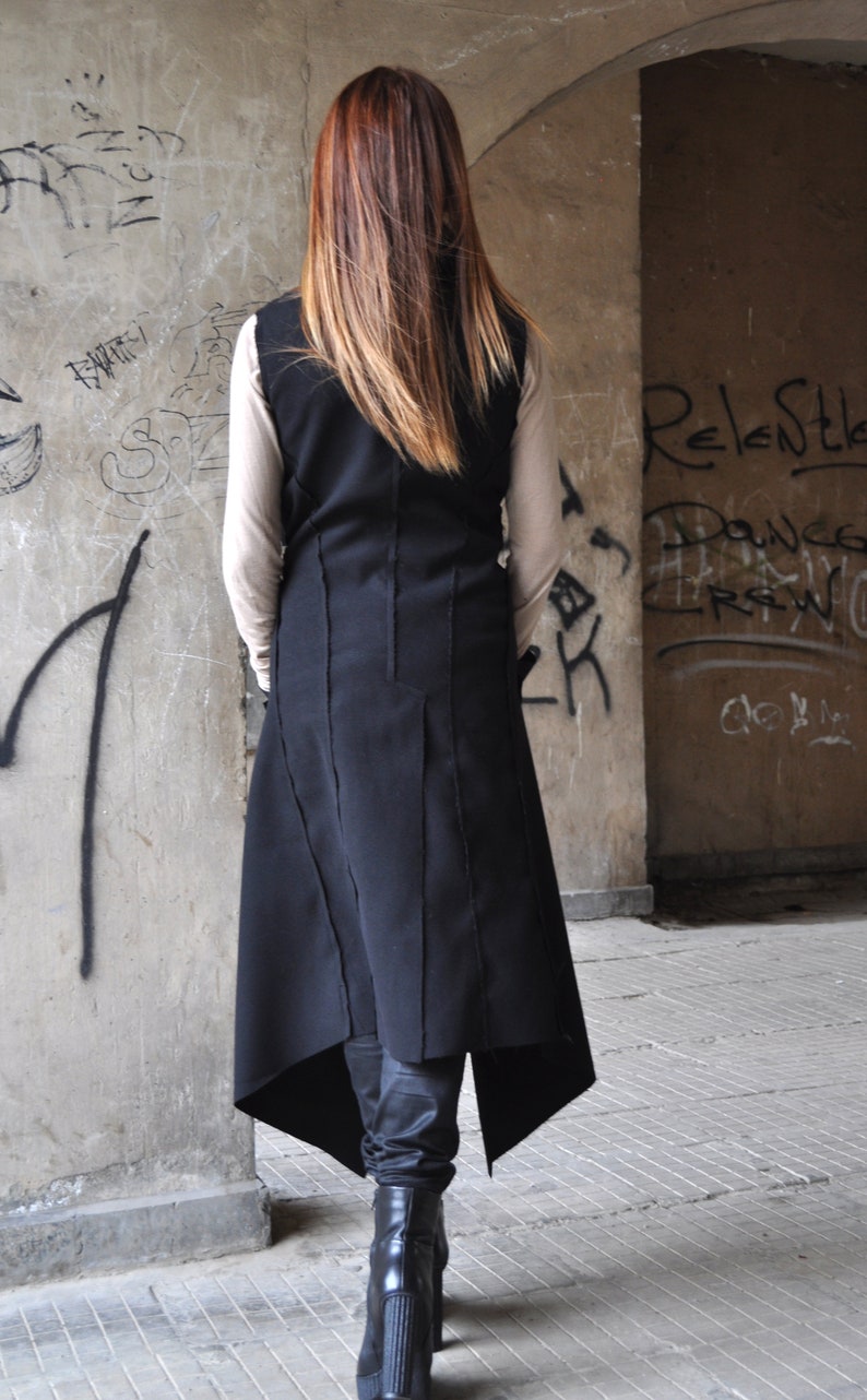 May include: A black asymmetrical vest with a long, straight cut and a high neckline. The vest is made of a thick, textured fabric and has a raw-edge finish. The model is wearing the vest over a white long-sleeved shirt and black pants.