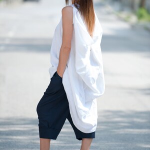 May include: A woman wearing a white linen top with a loose, flowing design and dark blue cropped pants. She is wearing white heeled sandals and sunglasses.