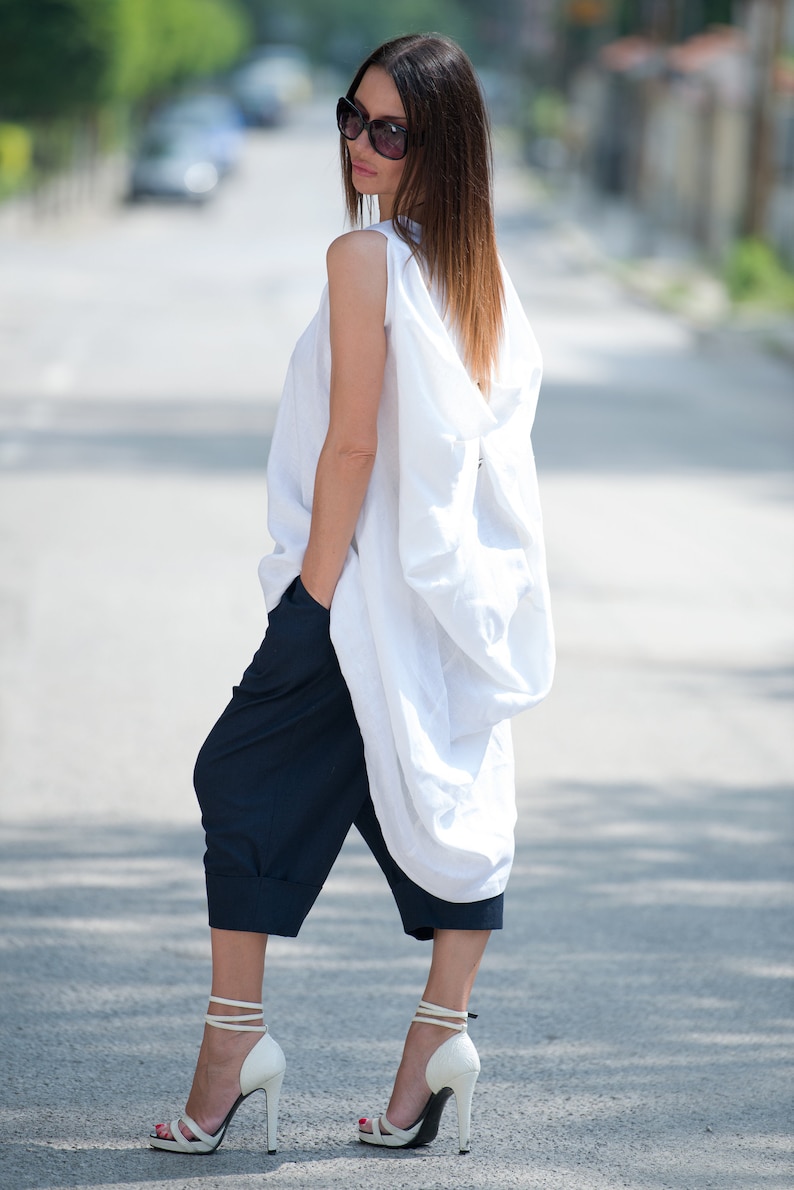 May include: A woman wearing a white linen top with a loose, flowing design and dark blue cropped pants. She is wearing white heeled sandals and sunglasses.