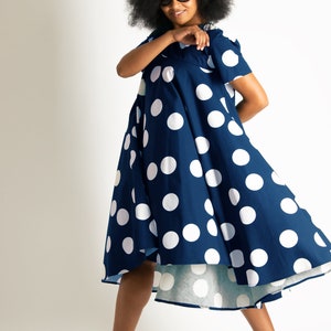 May include: A navy blue dress with large white polka dots. The dress has short sleeves and a flared skirt. The person in the image is wearing black high-heeled sandals and sunglasses. The background is plain white.