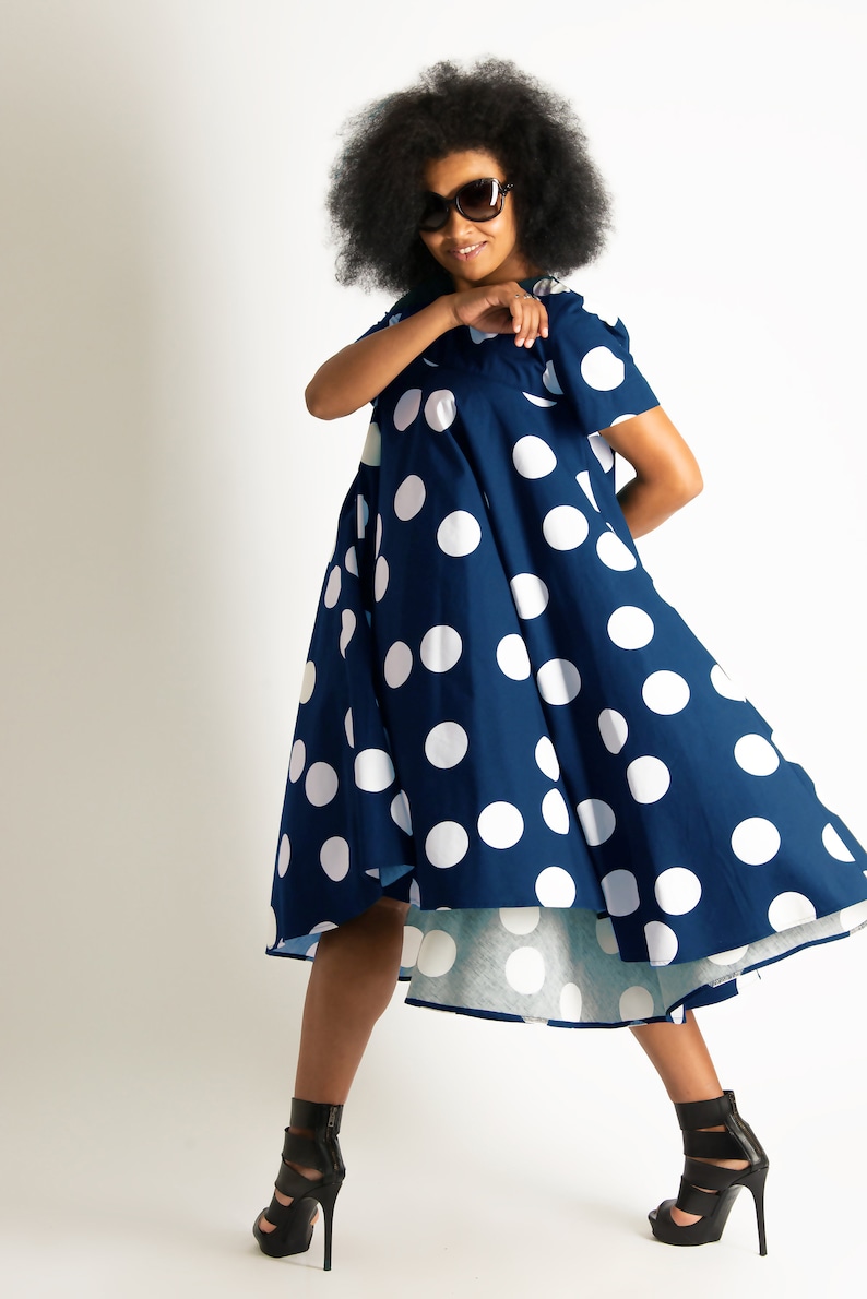 May include: A navy blue dress with large white polka dots. The dress has short sleeves and a flared skirt. The person in the image is wearing black high-heeled sandals and sunglasses. The background is plain white.
