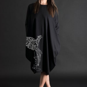 May include: A black, long-sleeved dress with a white parrot graphic and the word "Parrots" written in cursive on the side. The dress is loose-fitting and falls to the knees. The model is wearing black heels.