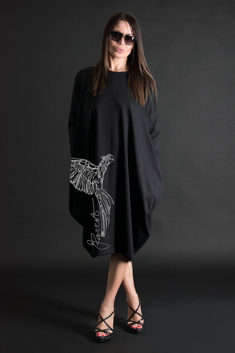 May include: A black, long-sleeved dress with a white parrot graphic and the word "Parrots" written in cursive on the side. The dress is loose-fitting and falls to the knees. The model is wearing black heels.
