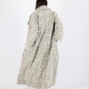 May include: A long, textured coat in shades of beige and gray, with a subtle pattern. The coat has a wide collar and long sleeves. The hemline reaches the ankles. The person is wearing patterned high heels.